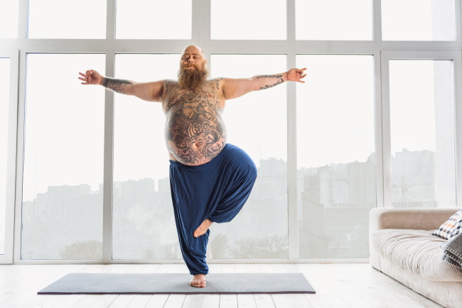 Gut beleibter Mann (Statur wie Buddha), nur mit Jogginghose bekleidet. Er hat einen langen Bart und der ganze Oberkörper ist tätowiert. Er steht in Yogapose auf einem Bein und hat die Augen geschlossen. Er macht einen sehr entspannten Eindruck. Gut beleibter Mann (Statur wie Buddha), nur mit Jogginghose bekleidet. Er hat einen langen Bart und der ganze Oberkörper ist tätowiert. Er steht in Yogapose auf einem Bein und hat die Augen geschlossen. Er macht einen sehr entspannten Eindruck.